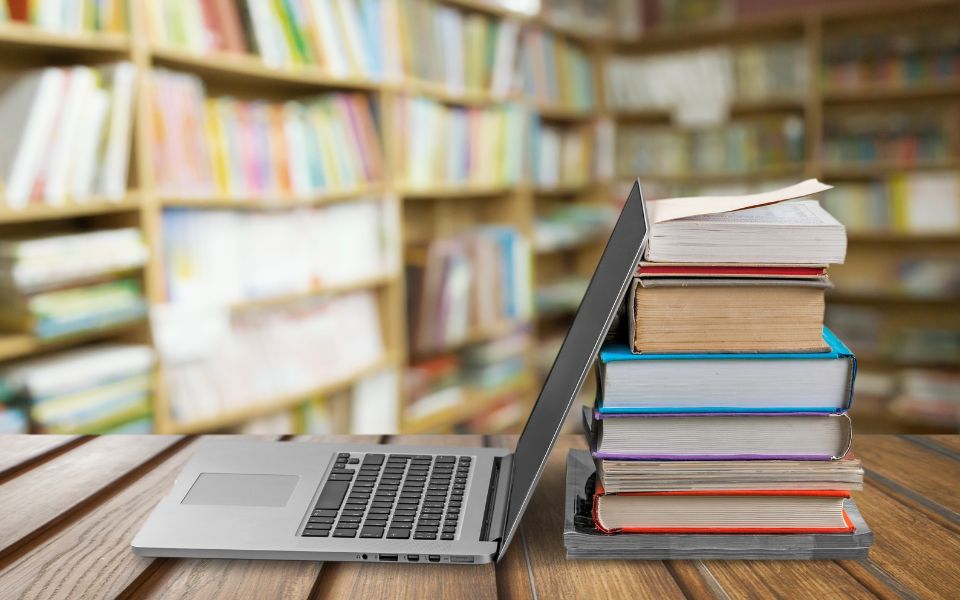 Fotografía de un ordenador portátil apoyado sobre una columna de libros. De fondo se ve una biblioteca