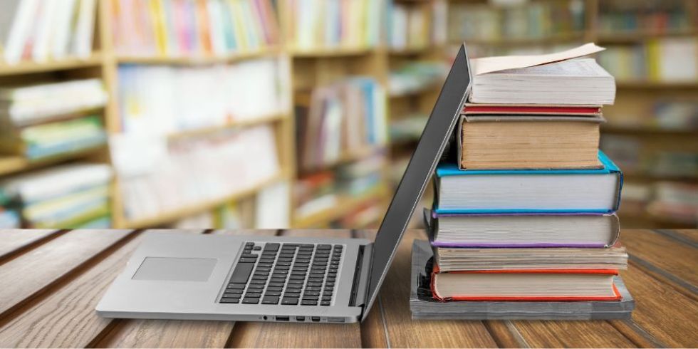 Un ordenador portátil sobre una mesa de madera y una pila de libros, dentro de una biblioteca, para acceder a la biblioteca virtual sobre autismo.