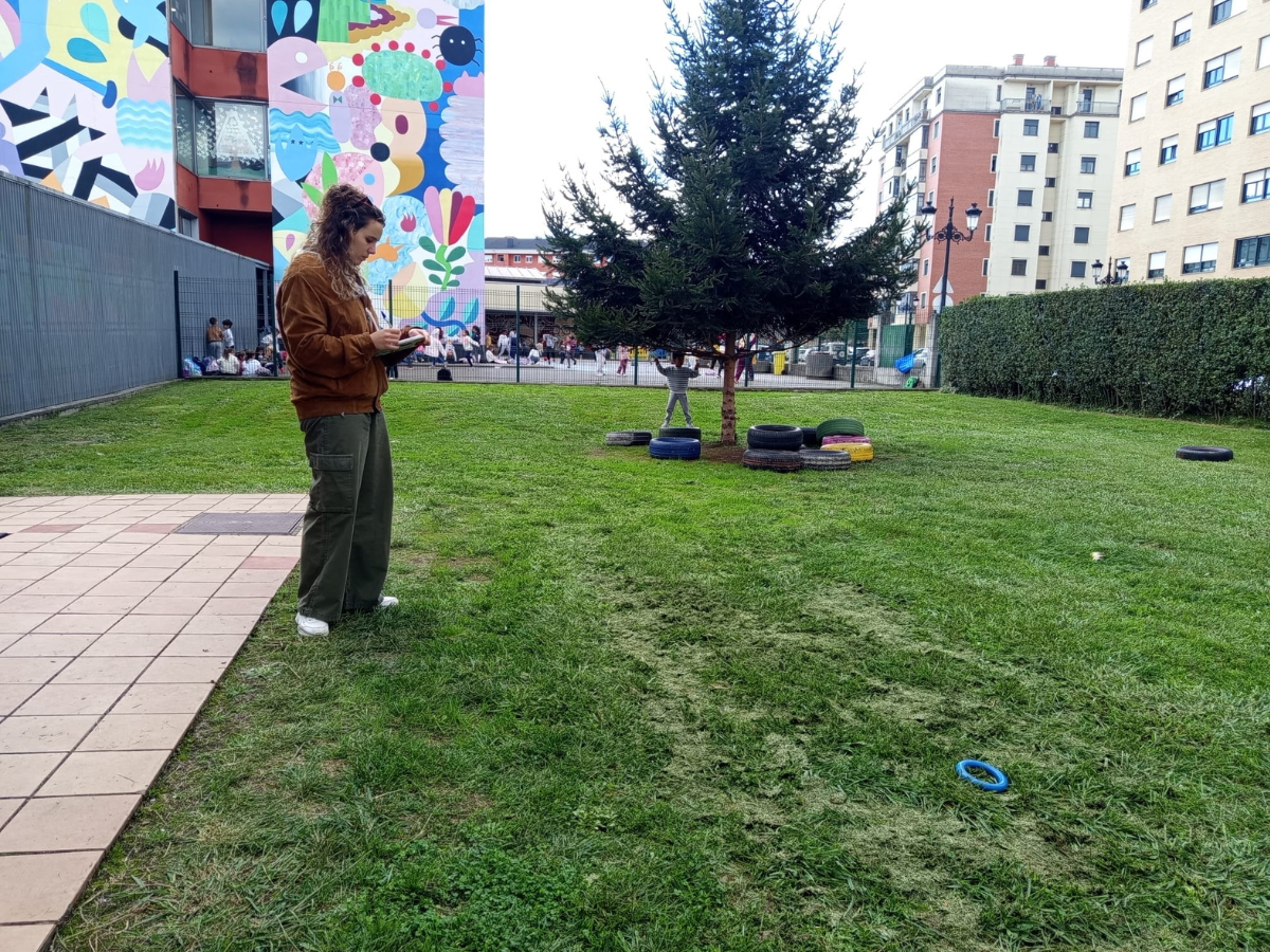 Una persona camina está de pie en un patio de colegio con mucho cesped y pintado donde hay una pared pintada de colores y está cogiendo notas en un cuaderno.